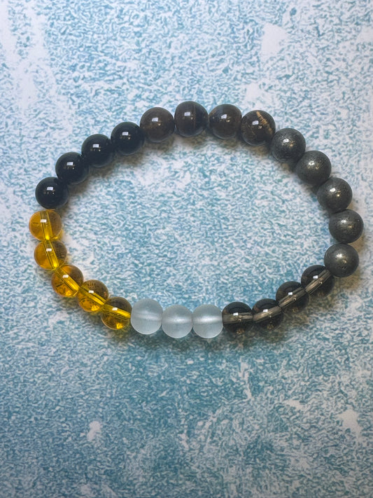 CraftnCrumble LLC's custom-made unisex bracelet, crafted from natural stones, showcases an artful arrangement of beads in yellow, clear, dark gray, and brown hues. This exquisite piece elegantly rests on a light blue textured surface.