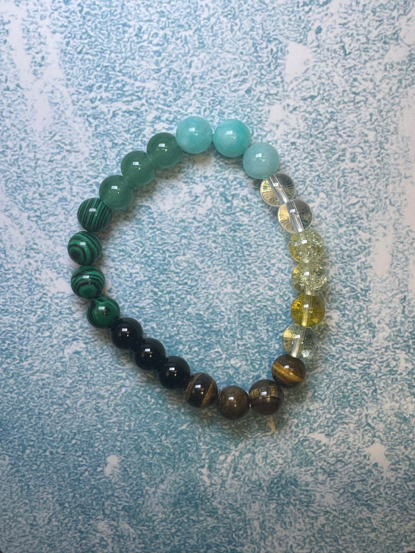 CraftnCrumble LLC's custom-made unisex bracelets crafted from natural stones feature a beautiful gradient of blue, green, brown, and yellow hues in a circular pattern set against a textured blue background. This versatile accessory is ideal for any occasion.