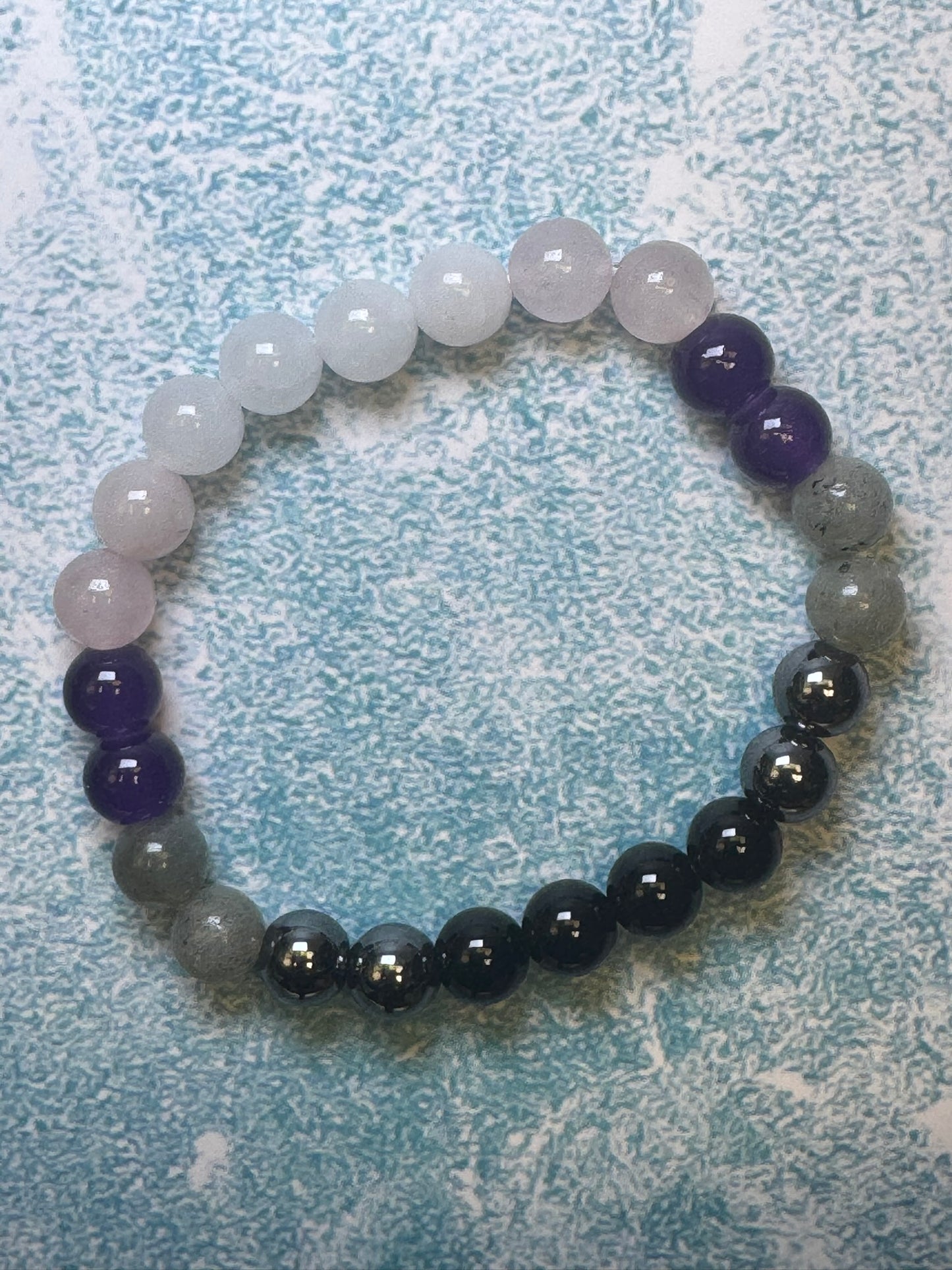 Experience the timeless appeal of a custom-made unisex bracelet crafted by CraftnCrumble LLC. This elegant bracelet features natural stones arranged in a circle, showcasing gradient colors that transition from white to dark gray with stunning purples and blues. The beautifully beaded design is displayed on a textured light blue surface.