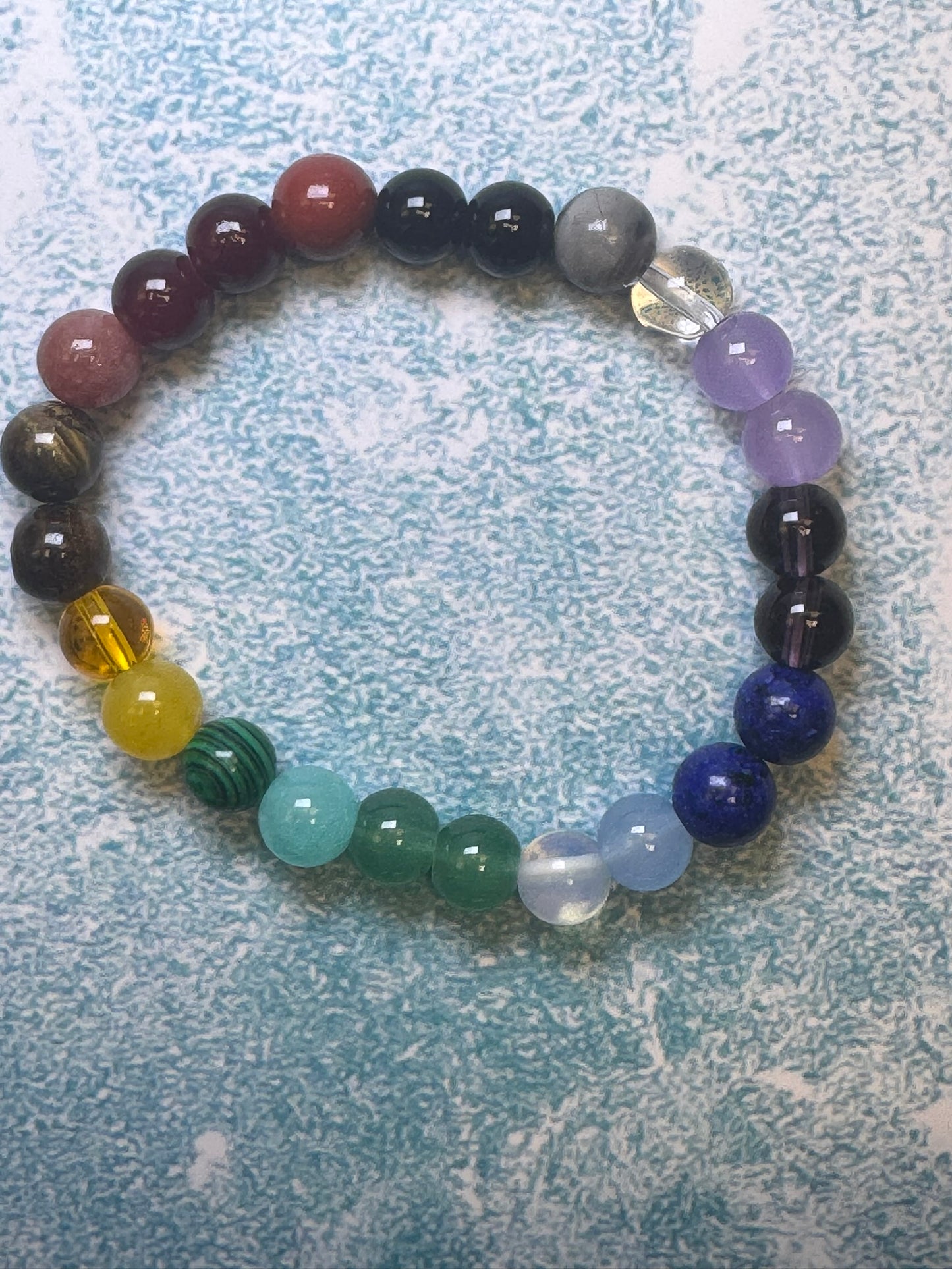 A custom-made bracelet by CraftnCrumble LLC, featuring multi-colored natural stone beads, elegantly rests in a circle on a textured blue and white surface. The unisex bracelet showcases an array of hues including red, pink, black, gray, purple, blue, green, yellow, and clear.