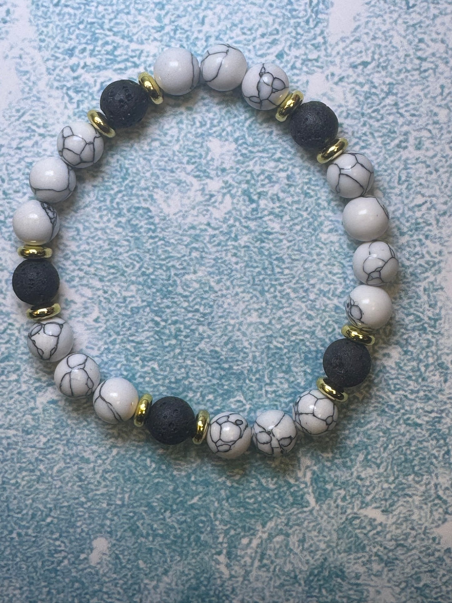 The CraftnCrumble LLC custom-made unisex bracelet, crafted from natural stones, elegantly showcases a mix of white beads with gray marbling, black beads, and small gold spacers against a textured blue background. This accessory effortlessly combines timeless style with the beauty of nature.