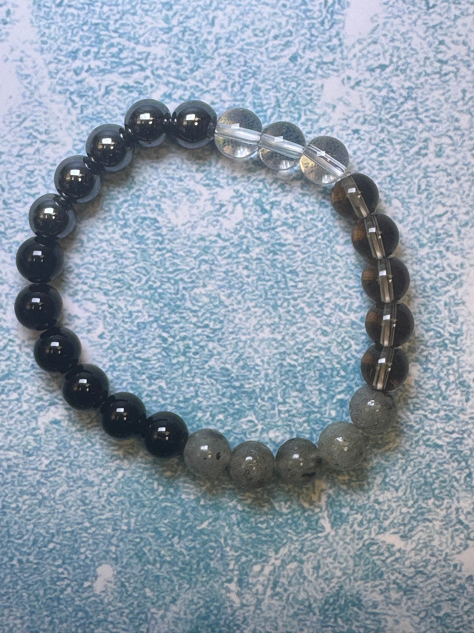 A custom-made unisex bracelet from CraftnCrumble LLC, crafted with alternating natural stones in black, transparent, and gray hues, is elegantly displayed on a textured light blue surface.