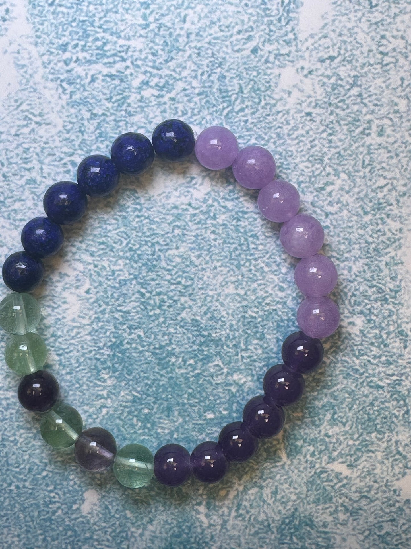 A Custom-made unisex bracelet from CraftnCrumble LLC, featuring alternating purple, green, and dark blue natural stones, forms a circle on a textured light blue surface. It's an ideal choice for those seeking bracelets that effortlessly combine elegance and style.