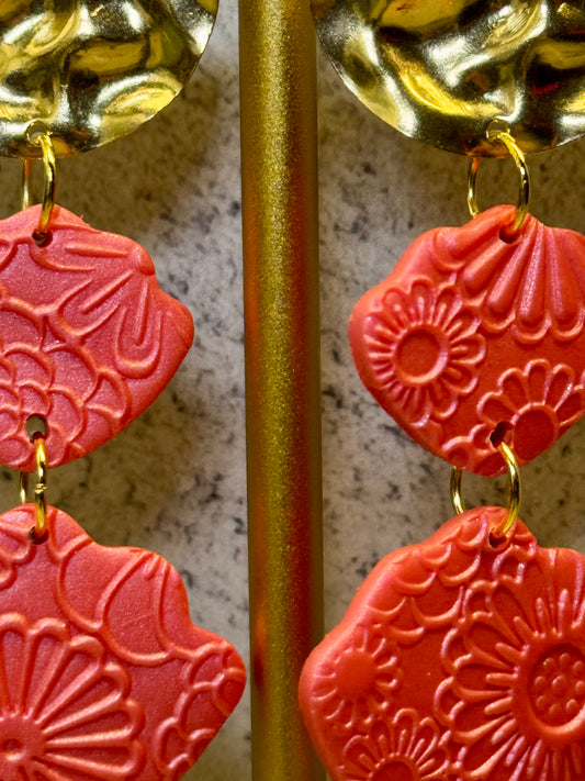 A close-up reveals the intricate details of CraftnCrumble LLC's Polymer Clay Chrome Earrings. These coral-colored earrings, featuring elegant floral patterns, hang gracefully from a metallic rod. The hypoallergenic design is enhanced by gold-toned connectors that create a striking contrast with the vibrant clay. The background showcases a textured, stone-like appearance.